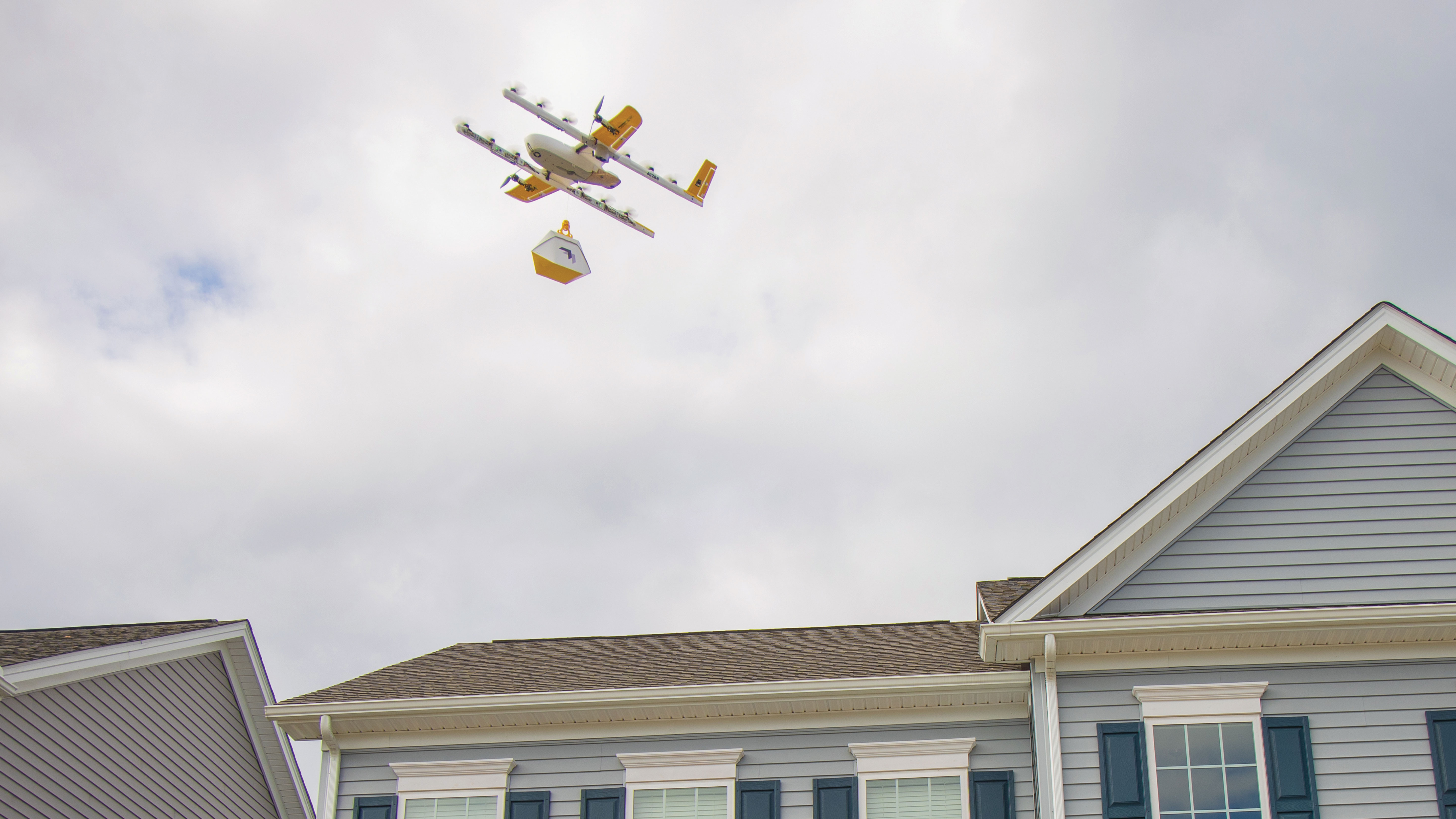 A delivery drone is flying above a suburban neighborhood, dropping a package onto the roof of a blue-grey house with white trim under a cloudy sky, illustrating modern delivery technology and residential homes.