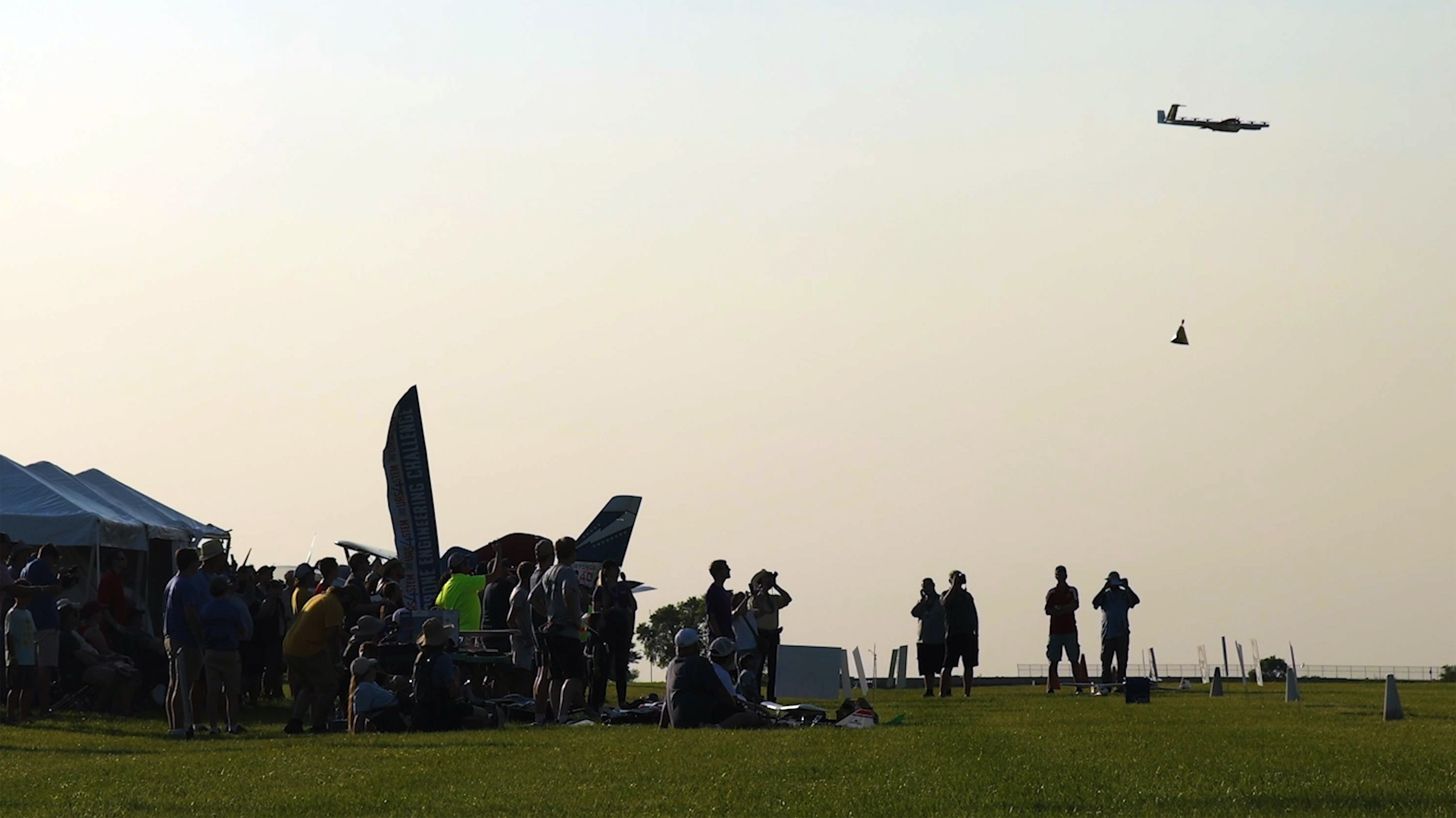 An airshow on a sunny day features a low-flying plane soaring over a crowd gathered on a green field, with tents and banners visible in the background.