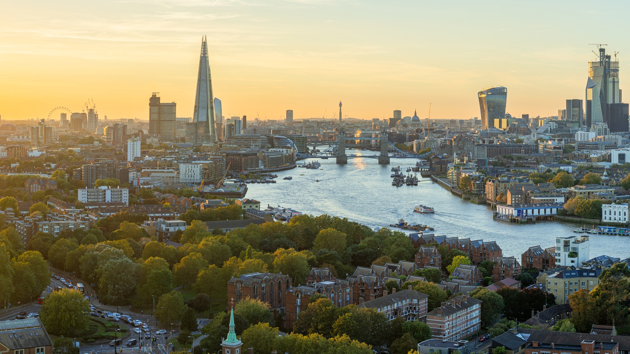 Aerial view of London at sunset featuring the River Thames, The Shard, and the distinctive modern architecture of the city skyline amidst lush green parks.