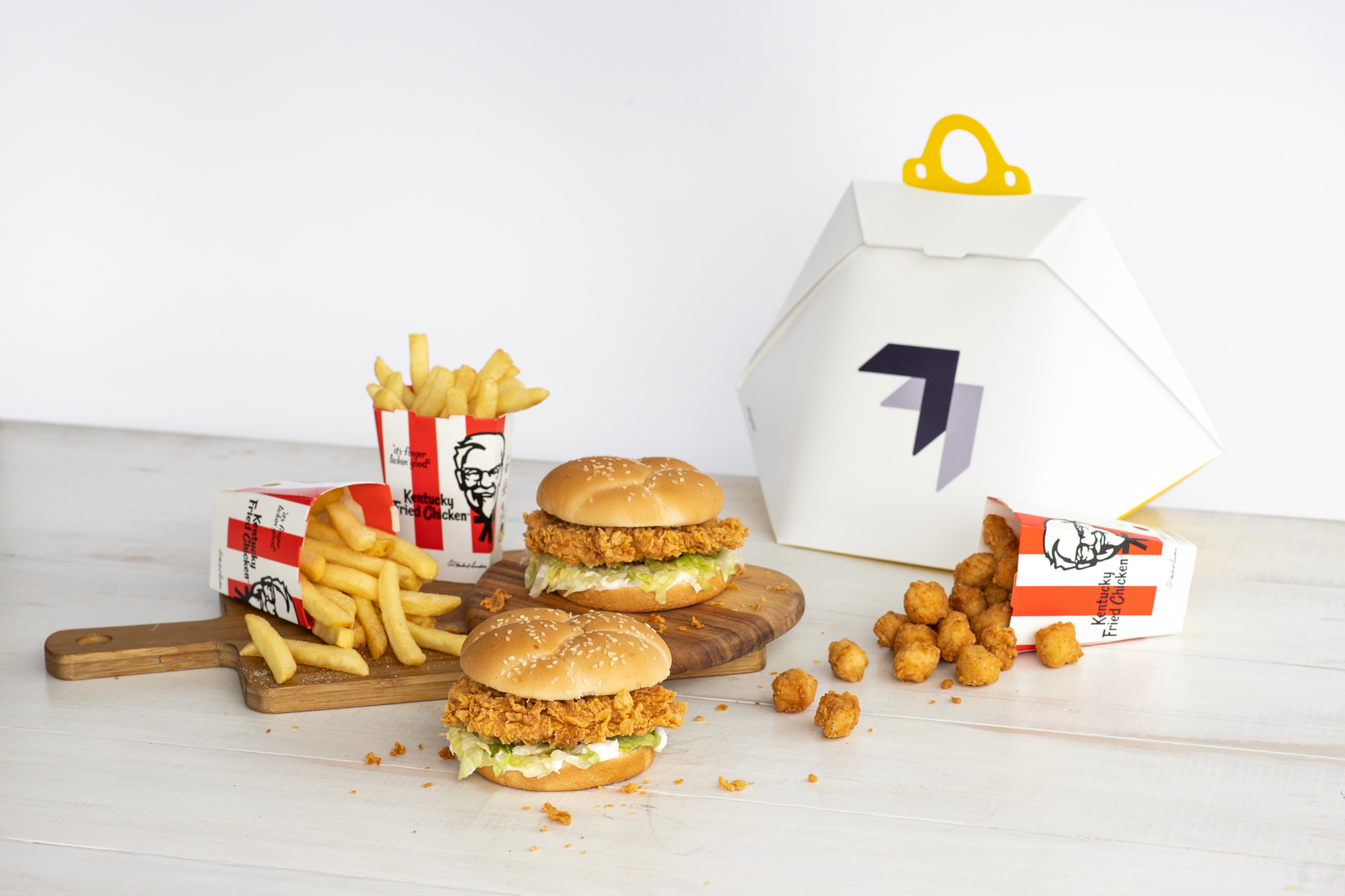 Two crispy chicken sandwiches with sesame seed buns are displayed on a wooden board, accompanied by servings of golden French fries and crunchy tater tots, placed alongside a branded takeout box.