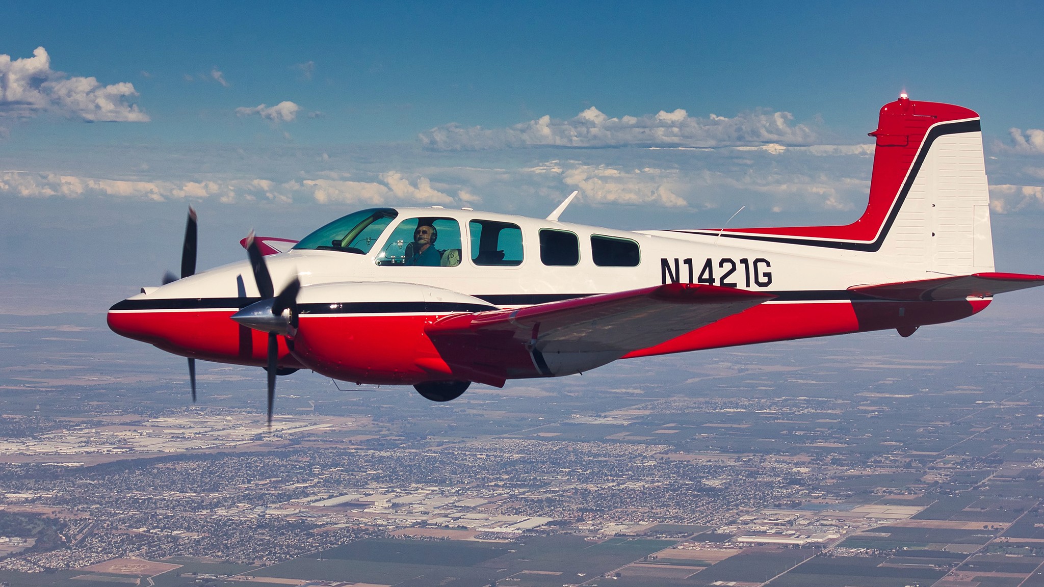 A sleek, red and white propeller airplane, identified by the number N1421G, is flying smoothly above a sprawling landscape under a clear blue sky with scattered clouds.