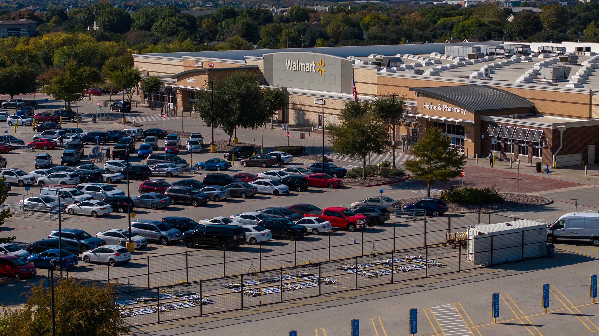 Aerial view of a busy Walmart parking lot filled with numerous parked cars, surrounded by green trees, with the store's signage visible on the large building on a clear day.