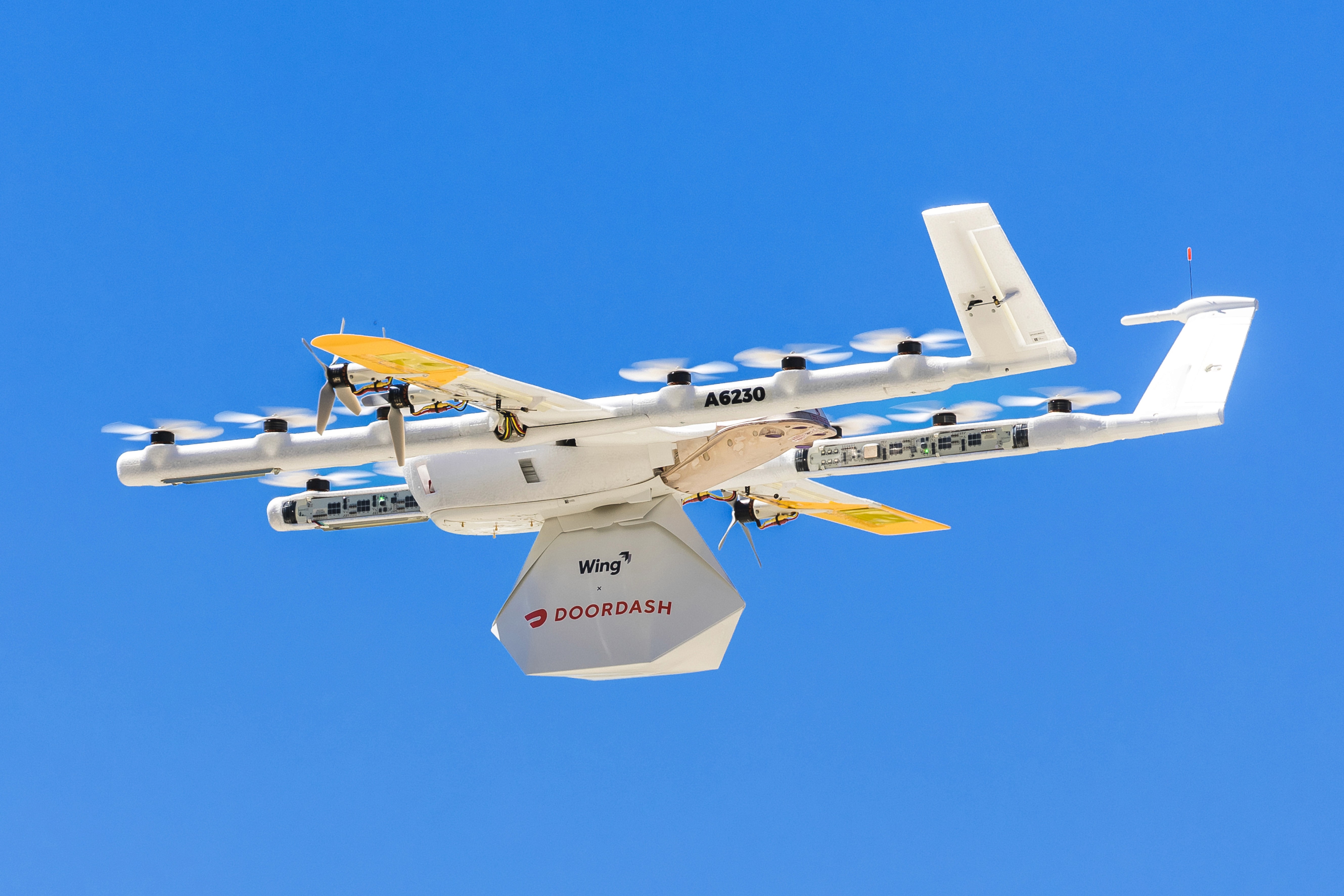 A white delivery drone with yellow accents and multiple propellers transports a package labeled "Wing" and "DoorDash" against a clear blue sky.