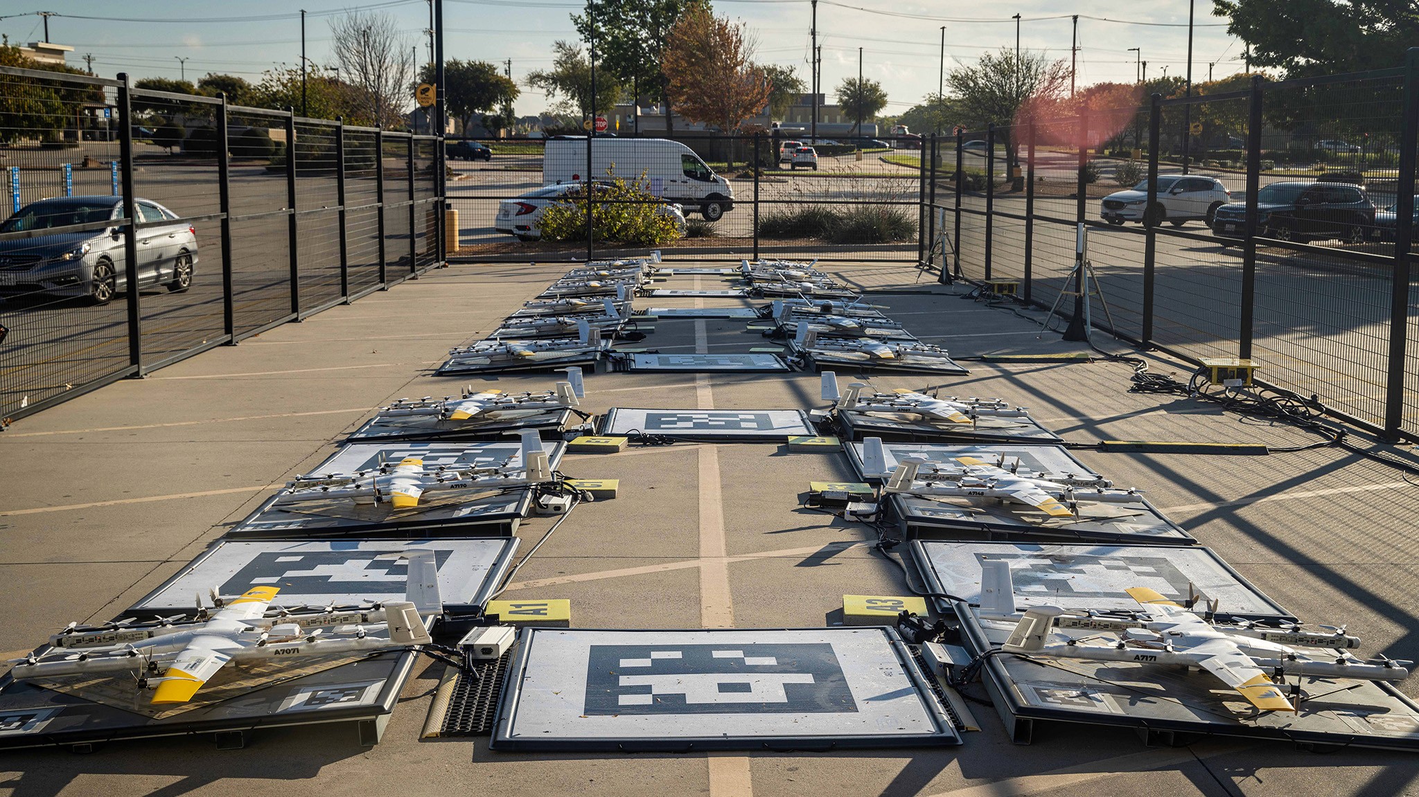 Four drone launching pads are aligned in two rows on a concrete area, each pad featuring a drone surrounded by safety fences, with trees, parked cars, and a white van visible in the background under a clear blue sky.