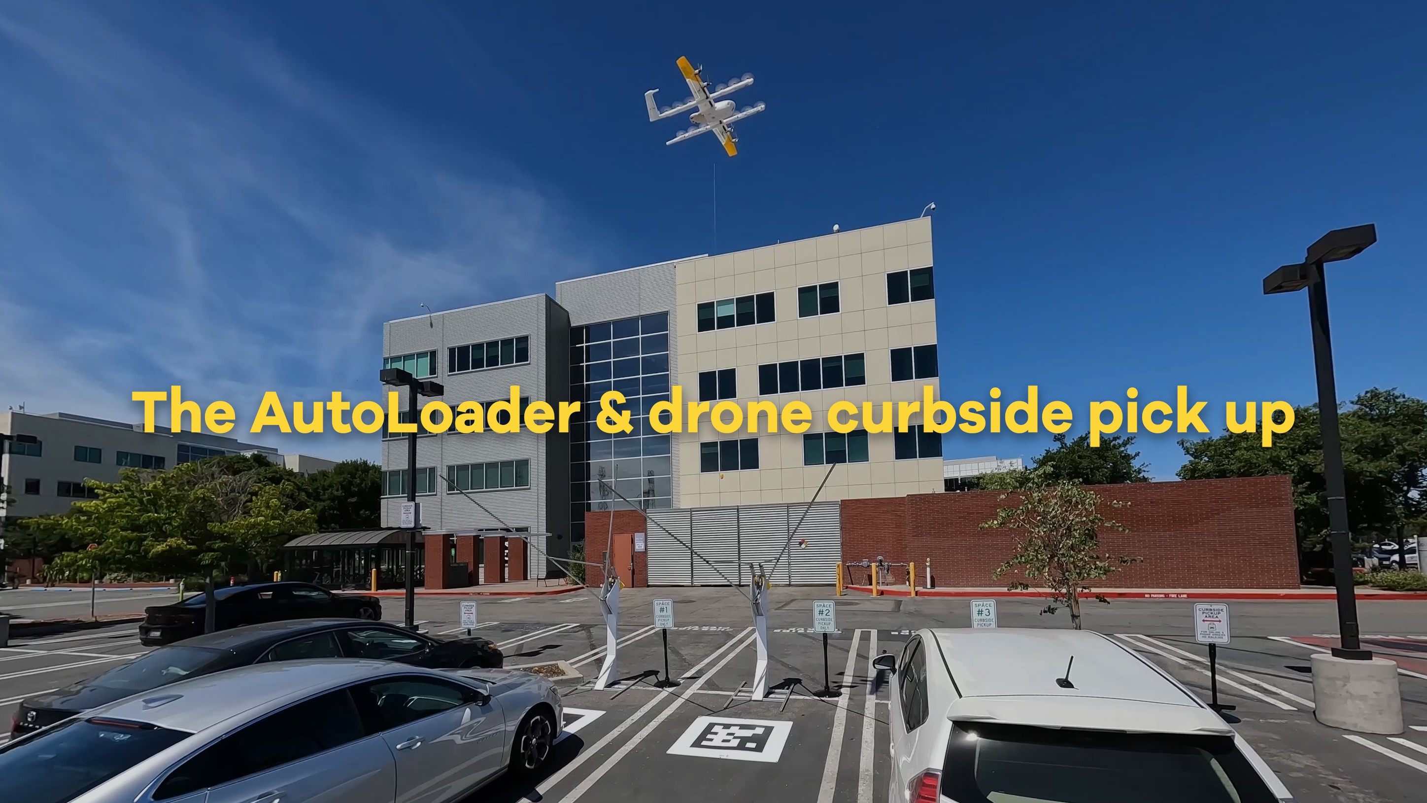 In an urban environment under a clear blue sky, a drone hovers above a modern multi-story building next to a nearly empty parking lot, highlighted by the caption "The AutoLoader & drone curbside pick up."