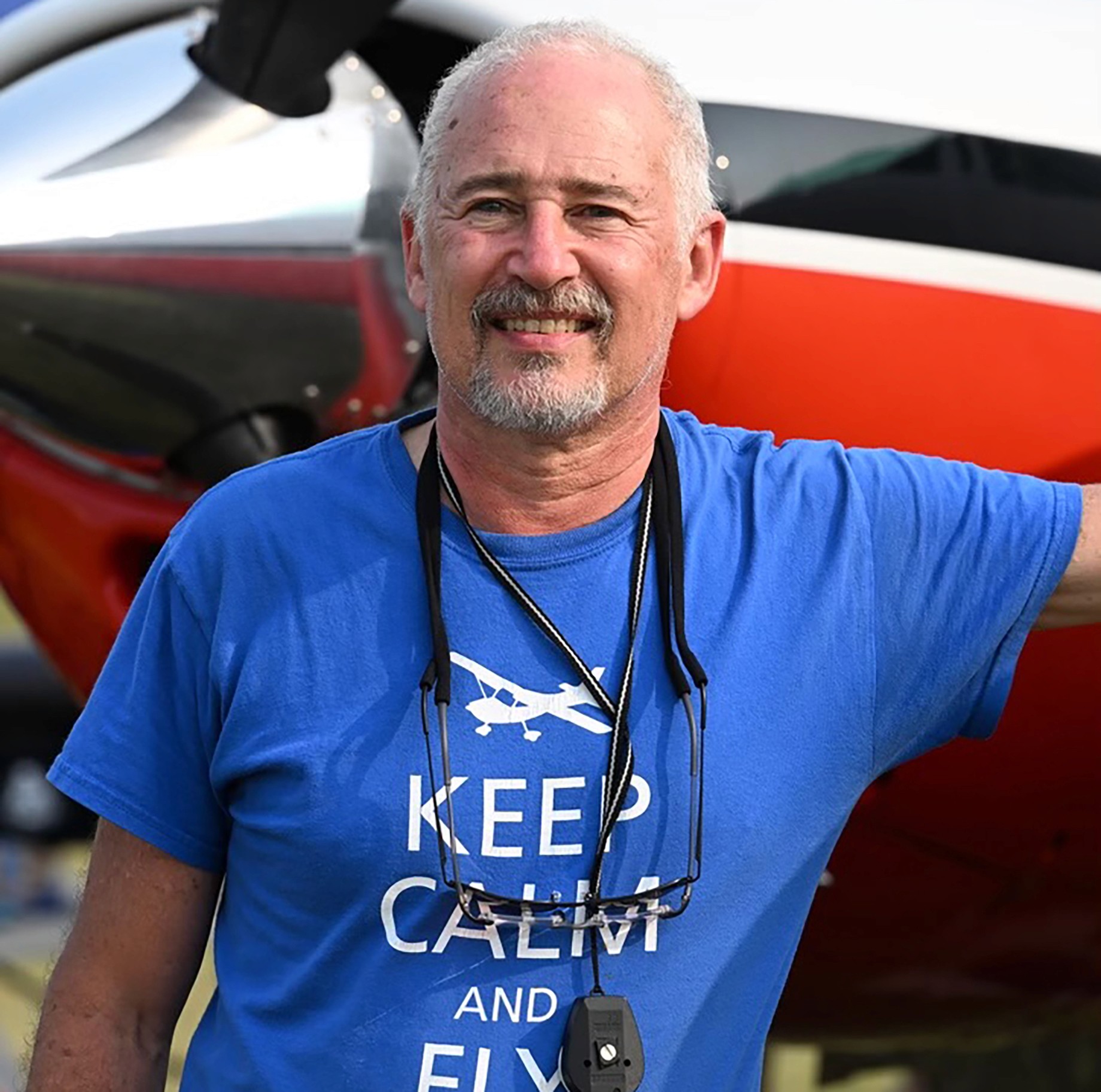 A man wearing a blue t-shirt with "Keep Calm and Fly" text and an airplane graphic, smiles while standing in front of a vibrant red and white aircraft, with the airplane's propeller partially visible in the background.