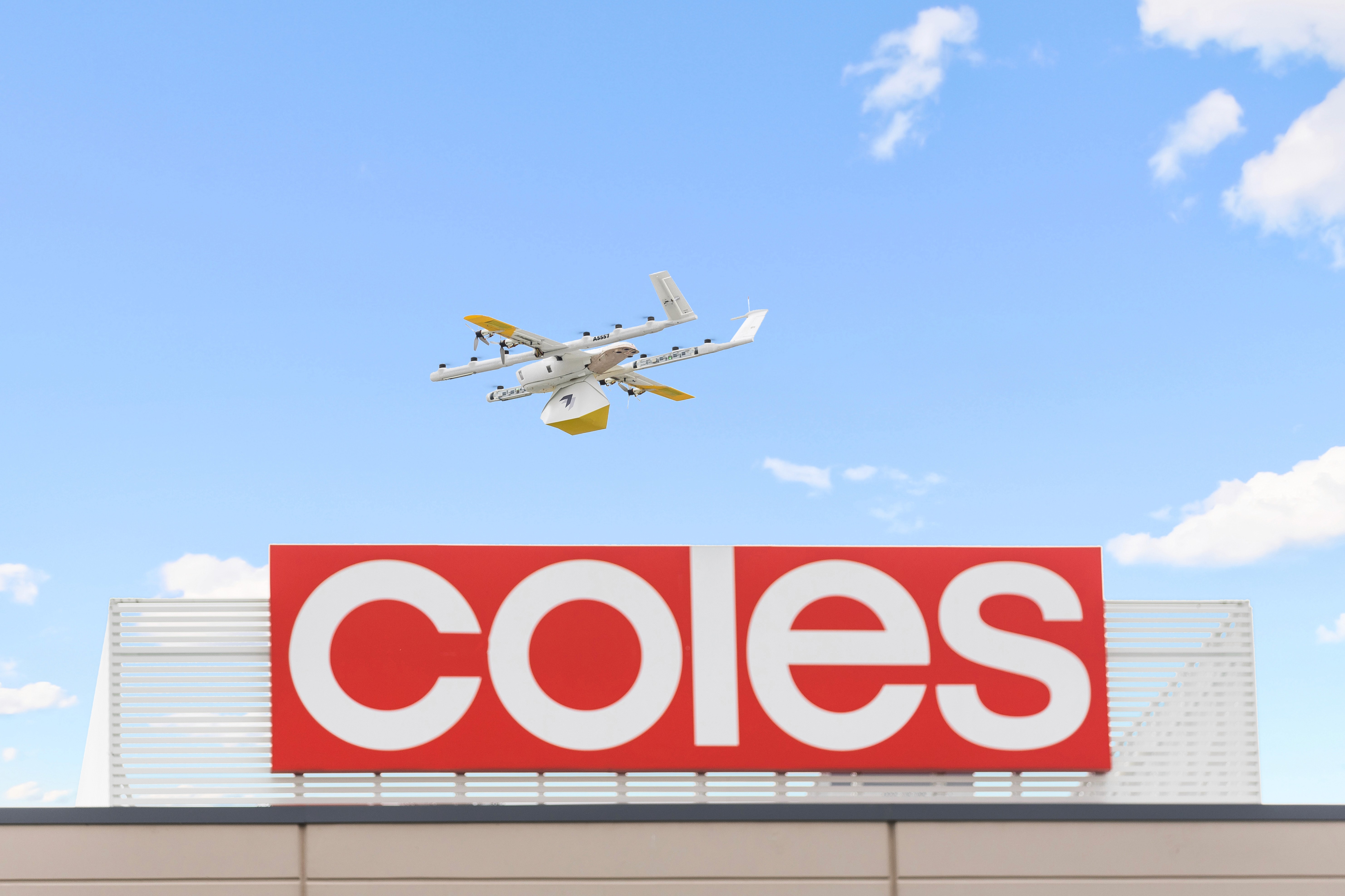 A drone with a sleek white and yellow design flies above a Coles supermarket sign against a backdrop of a clear blue sky scattered with fluffy white clouds.
