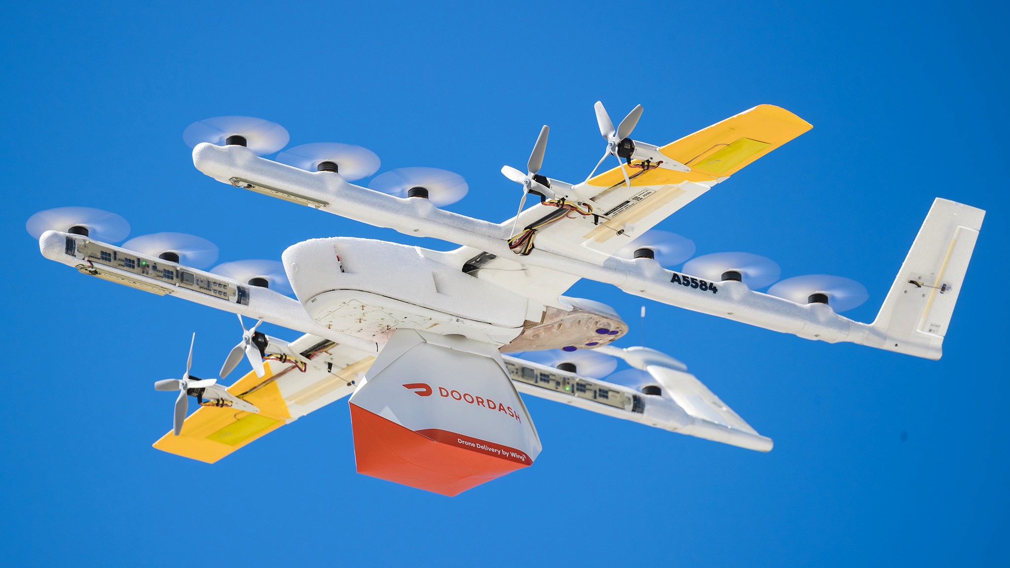A white and yellow drone in flight against a clear blue sky; it is carrying a red and orange package, demonstrating modern delivery technology.