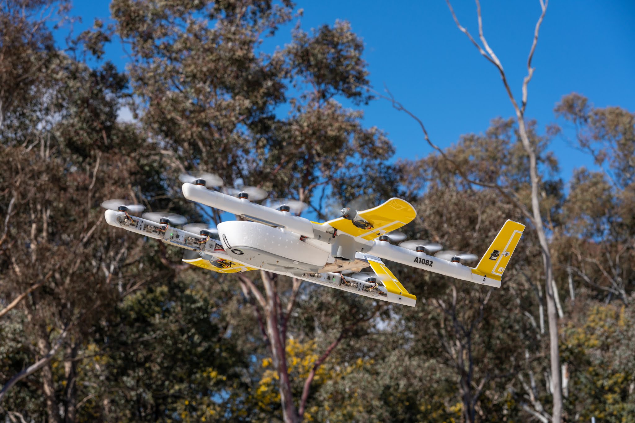 A white and yellow delivery drone with multiple propellers flies through a clear blue sky, surrounded by tall trees, demonstrating advanced aerial technology and innovative transportation solutions.