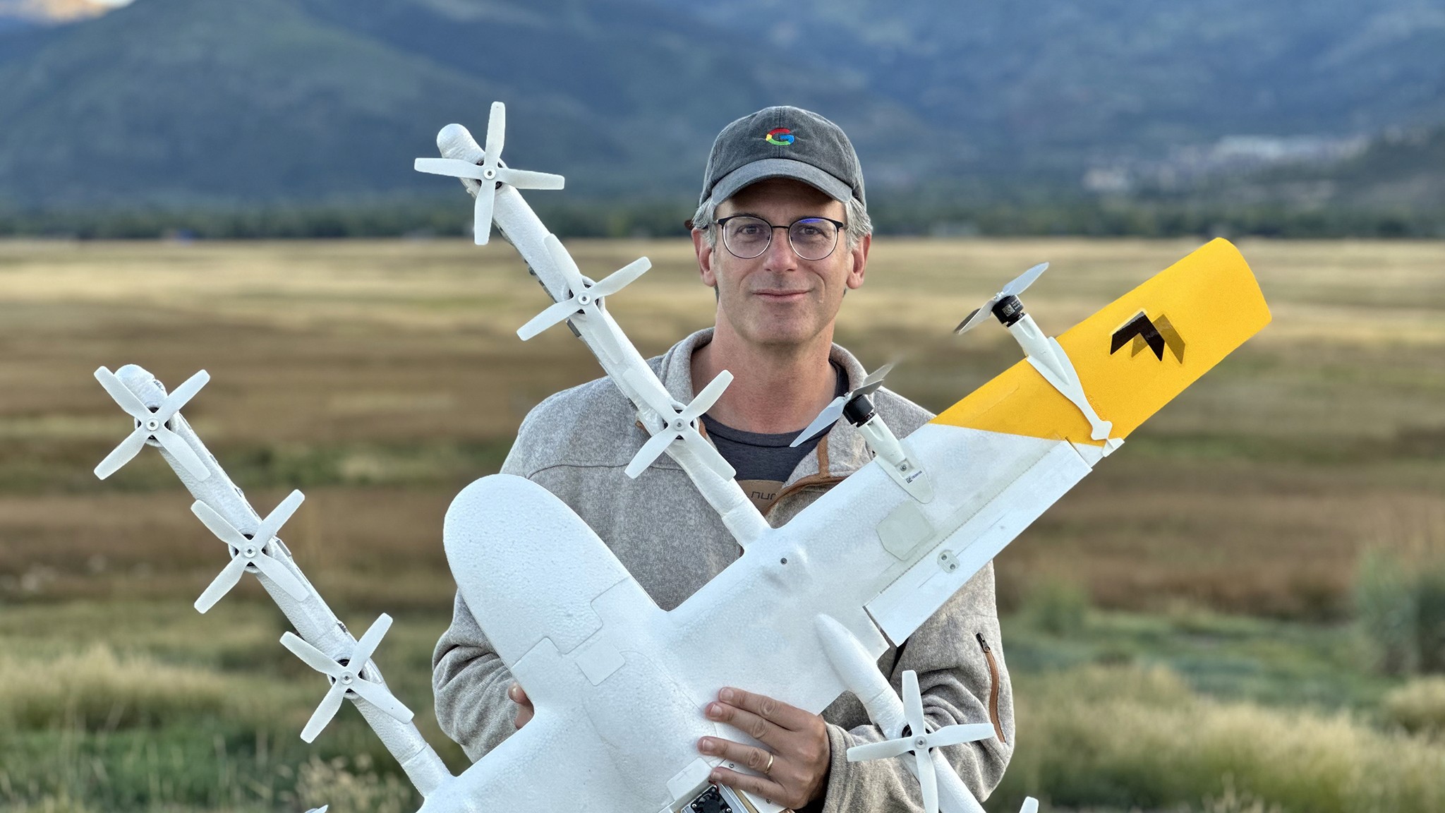 A person stands outdoors in a grassy field holding a white drone with multiple propellers, against a backdrop of rolling hills and a cloudy sky.