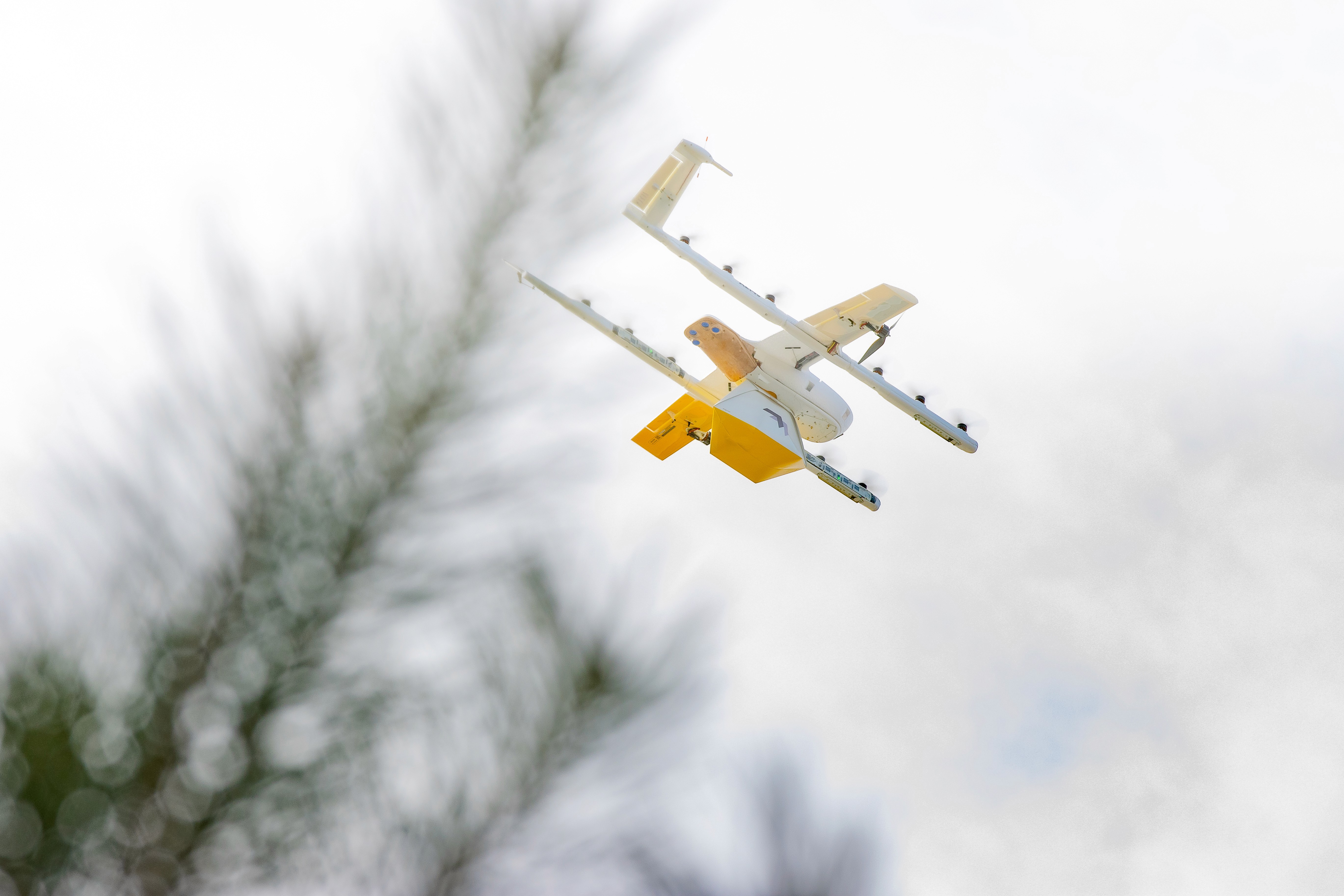 A delivery drone, equipped with a camera, flies through a clear blue sky while carrying a yellow package, surrounded by leafy green branches.