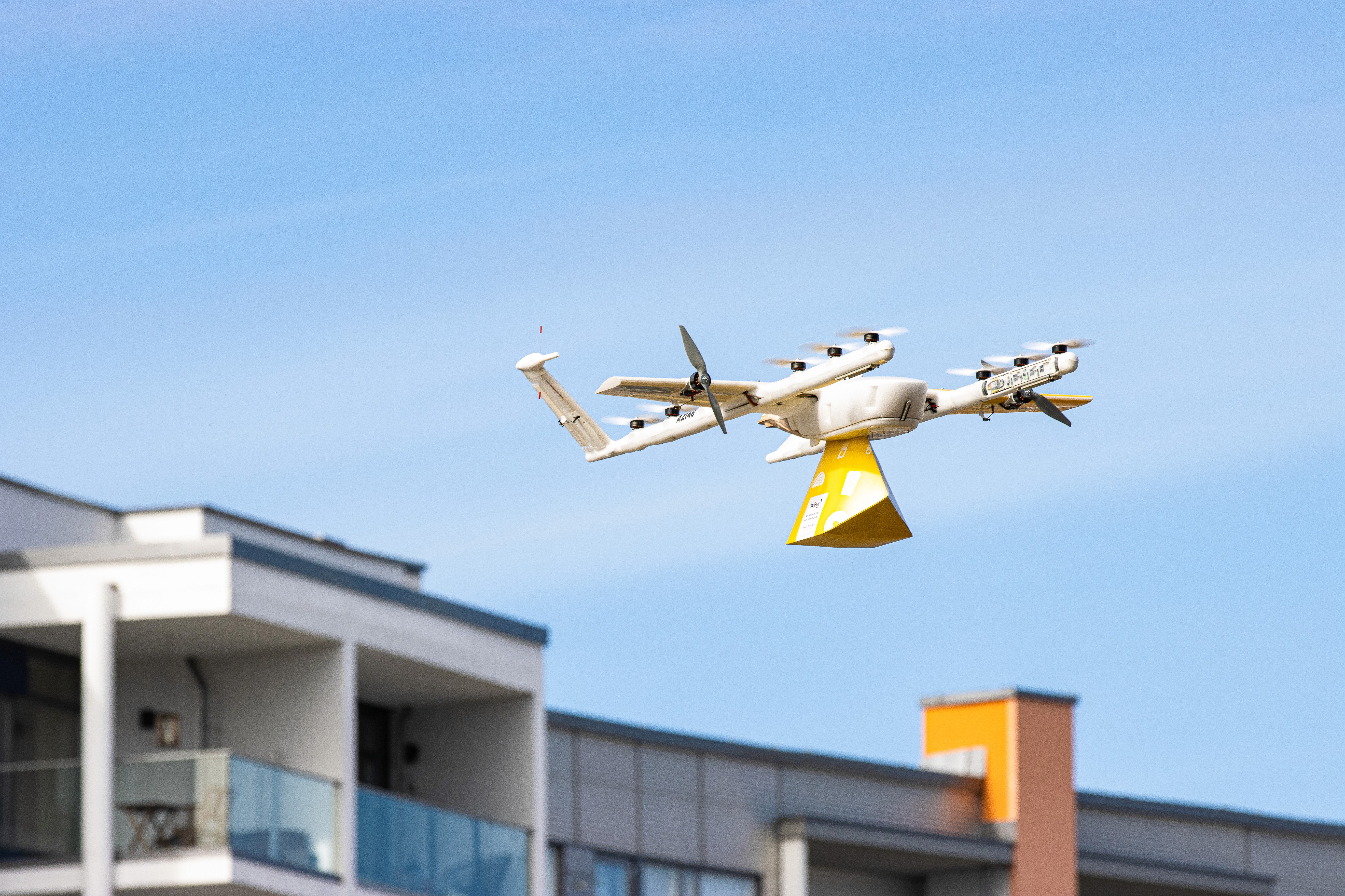 A white delivery drone with multiple rotors offers a package over a modern urban residential area, contrasting against a clear blue sky.