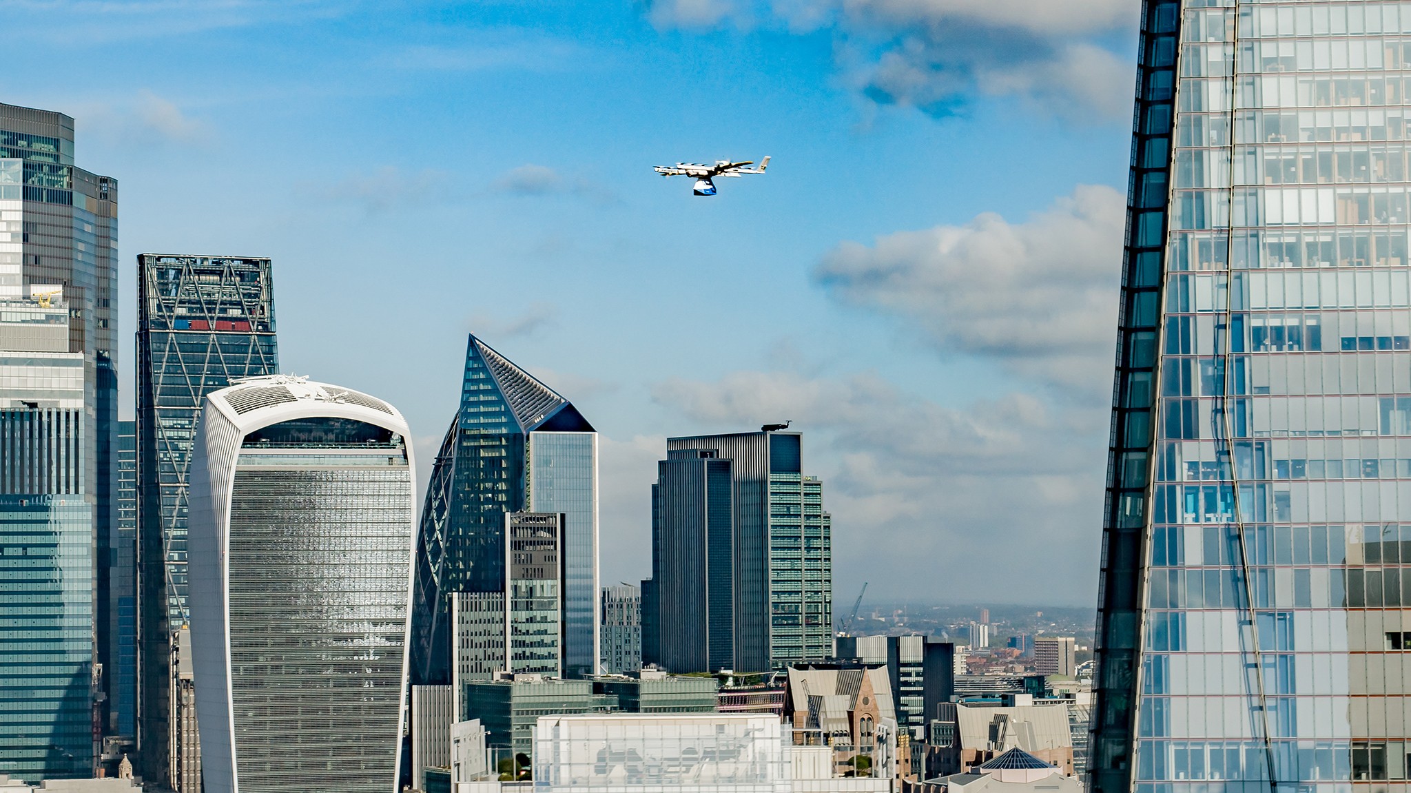 A white drone flies over the prominent skyline of a modern city, with distinctive skyscrapers like the Walkie Talkie building and the Leadenhall Building set against a clear blue sky.