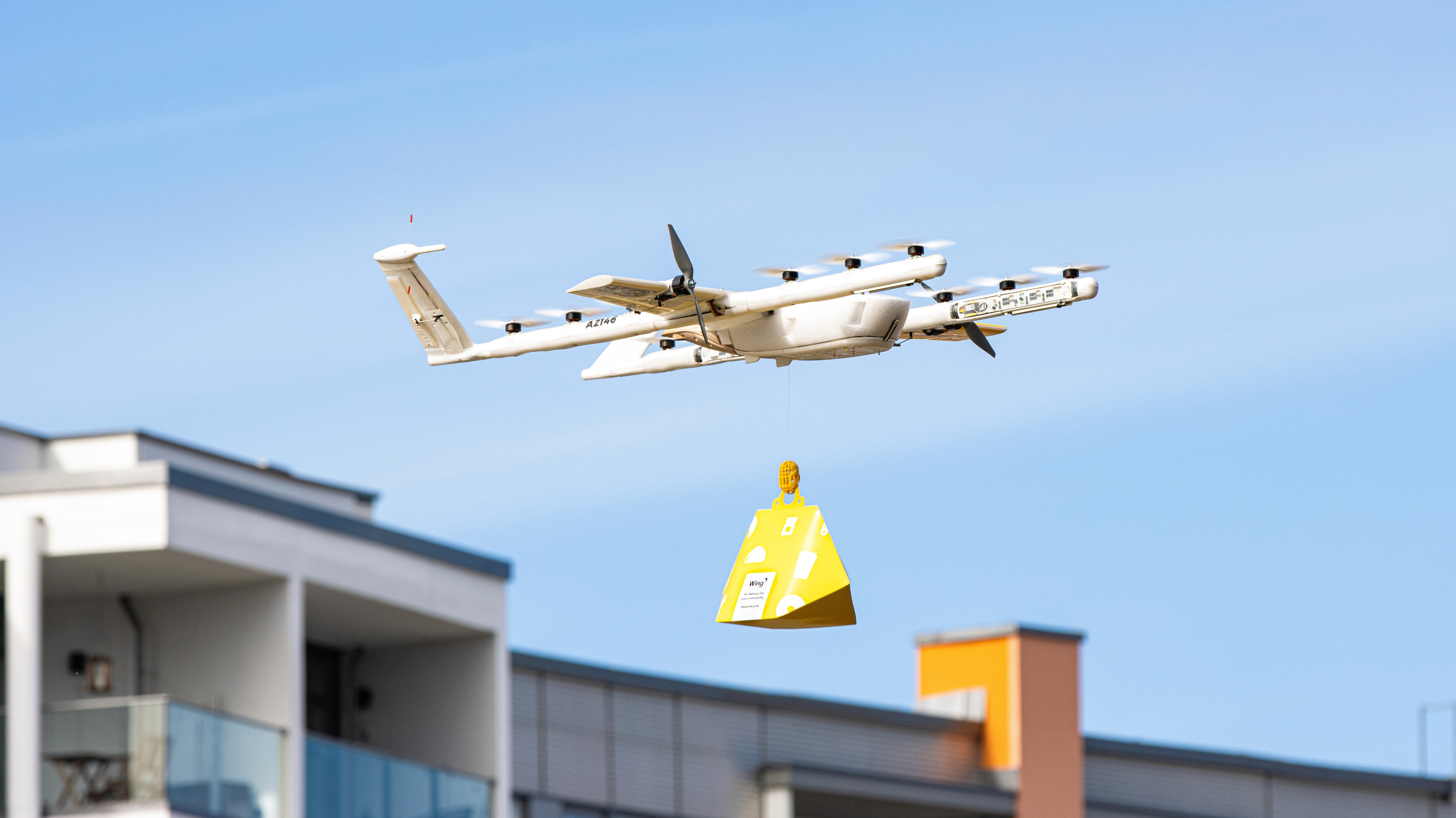 A white delivery drone flies above urban apartments, carrying a yellow parcel beneath a clear blue sky.