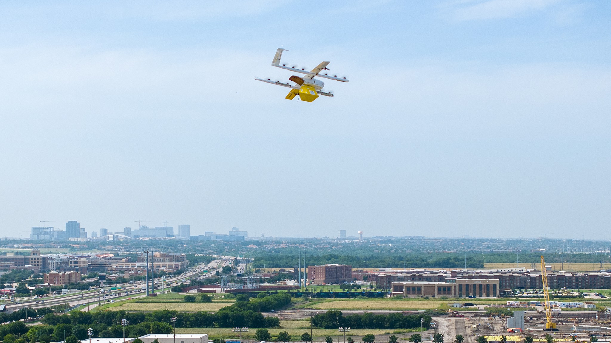 A white drone with yellow accents flies over an urban landscape with sprawling roads and distant city buildings under a clear blue sky.