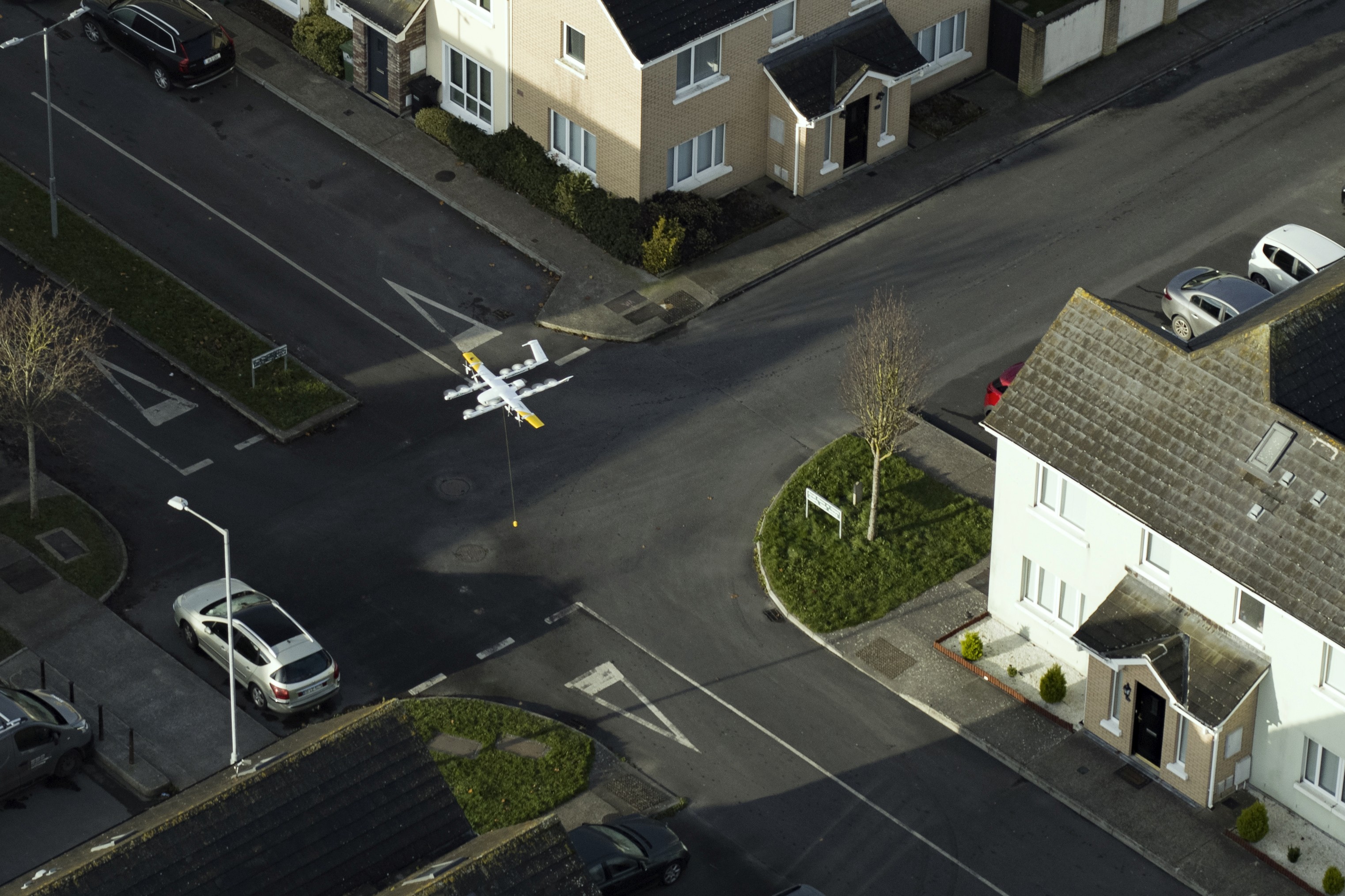 A small drone flies over a residential neighborhood intersection, surrounded by parked cars and houses, showcasing modern drone technology in urban delivery.