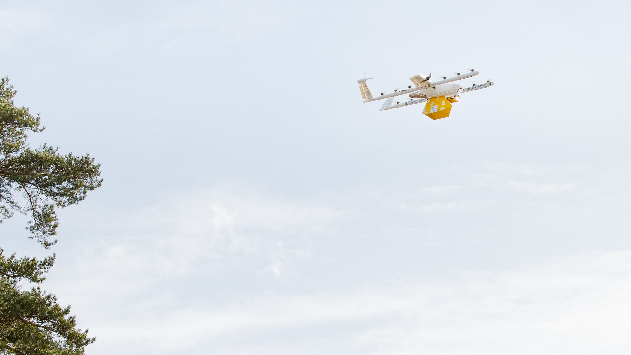 A white and yellow drone in flight against a clear blue sky; it is carrying a red and orange package, demonstrating modern delivery technology.