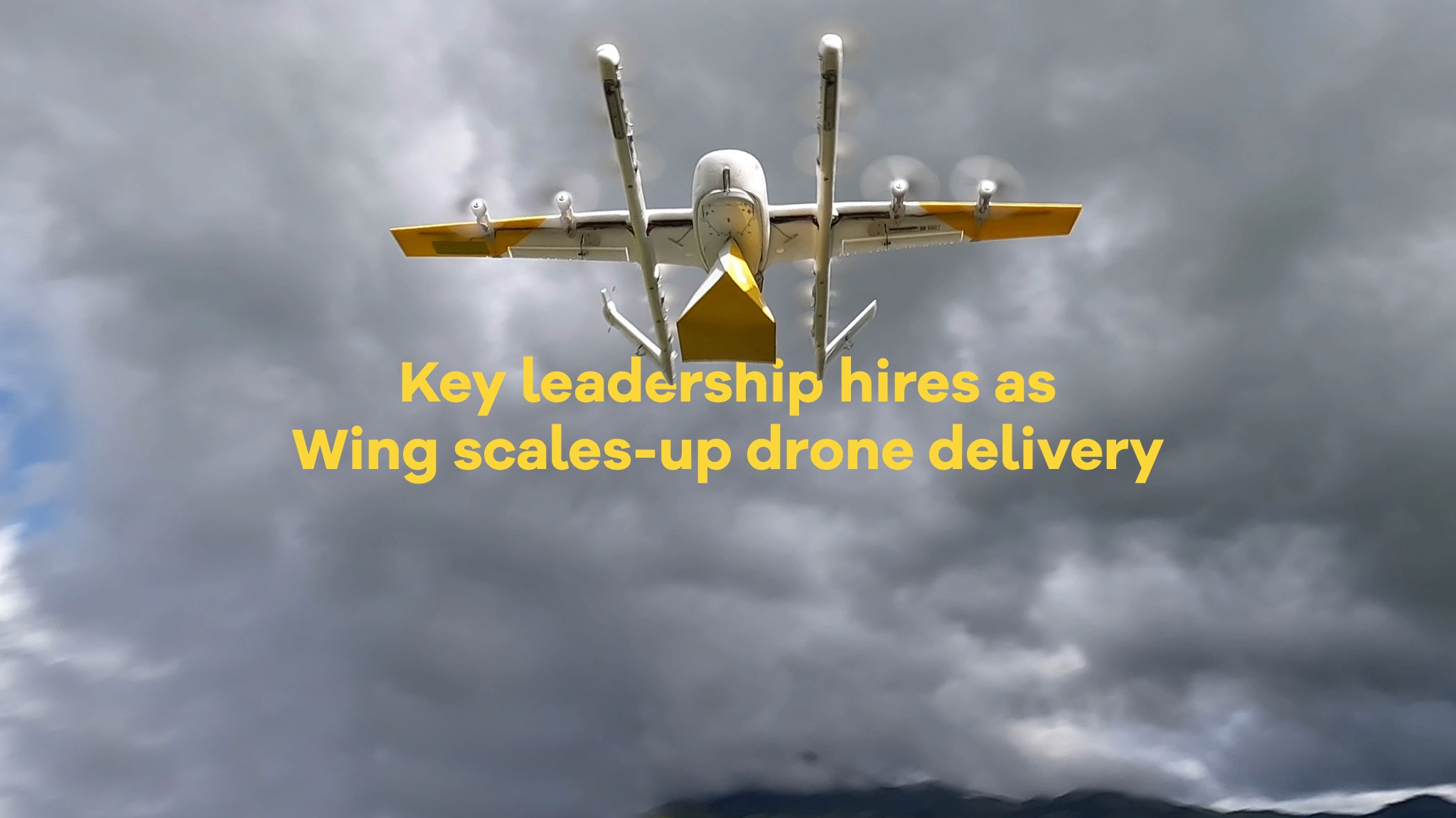 An advanced drone with yellow and white livery flies under a cloudy sky, illustrating Wing's focus on drone delivery expansion.