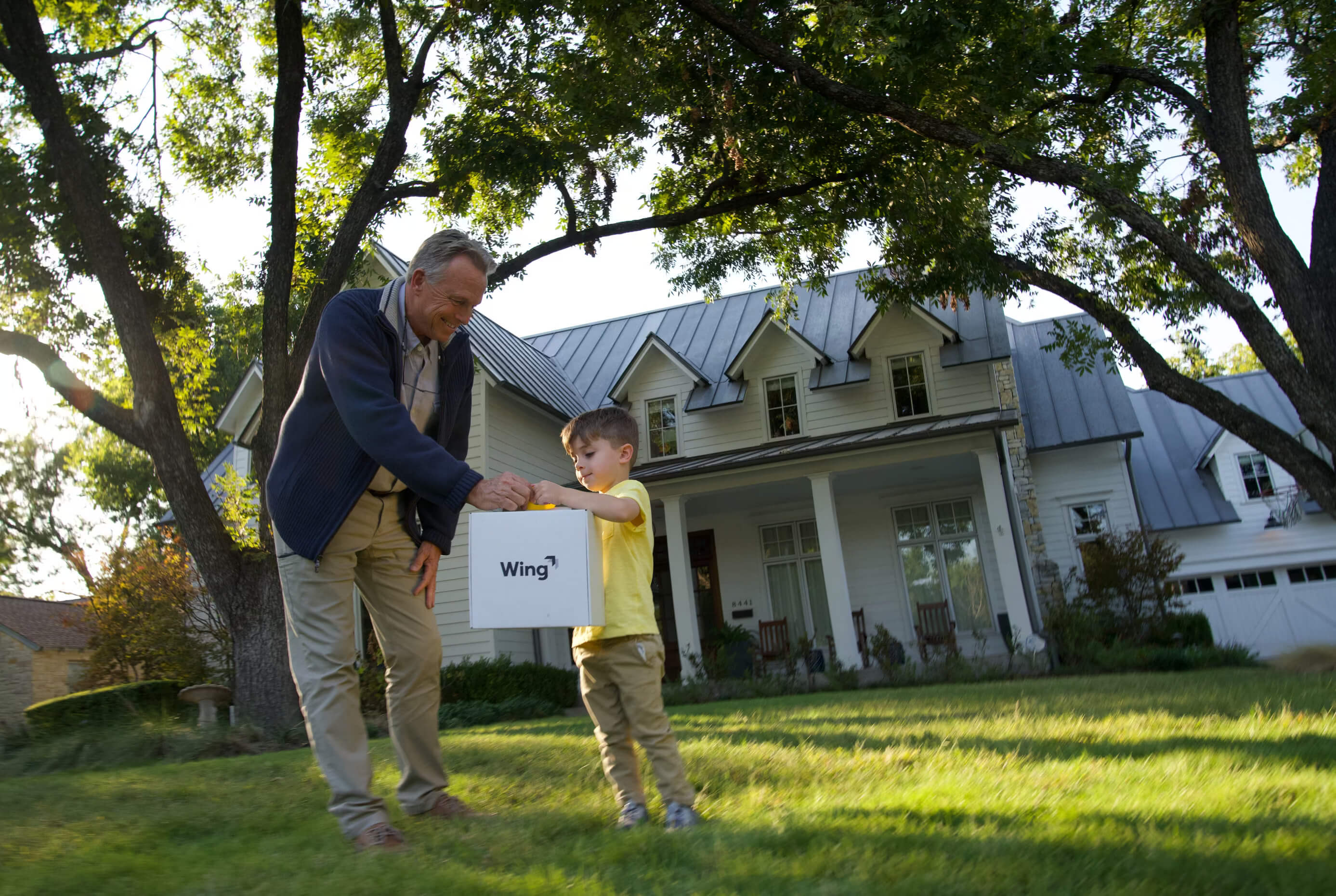 A grandfather and child stand on a lush green lawn in front of a large, charming house with tall trees, as they receive a drone  中国168体彩网-实时官方认证 Delivery package marked "Wing".