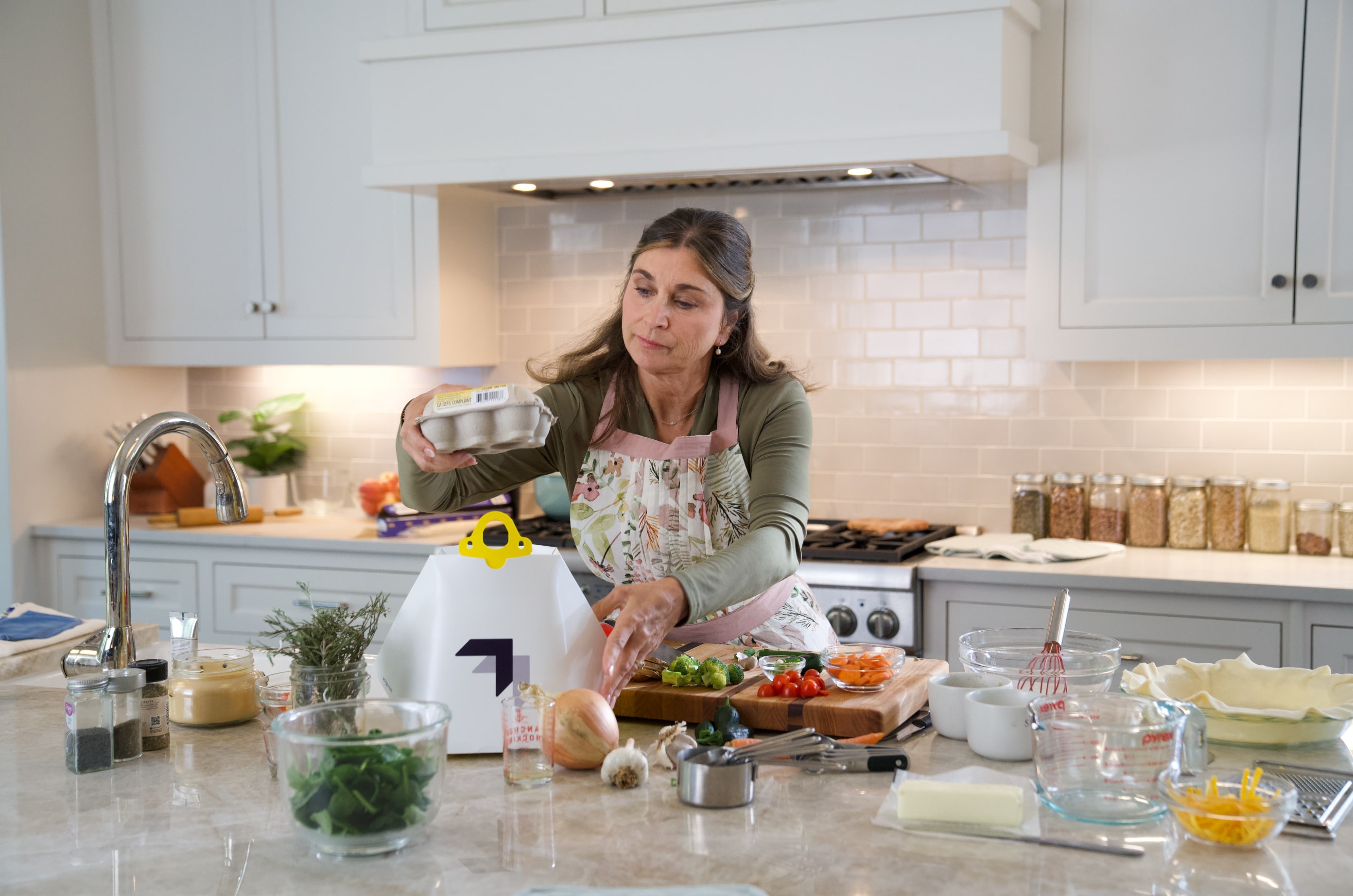 A woman in a kitchen prepares a meal, surrounded by fresh vegetables, spices, and cooking utensils, while wearing an apron amidst a backdrop of white cabinets and a tiled backsplash.