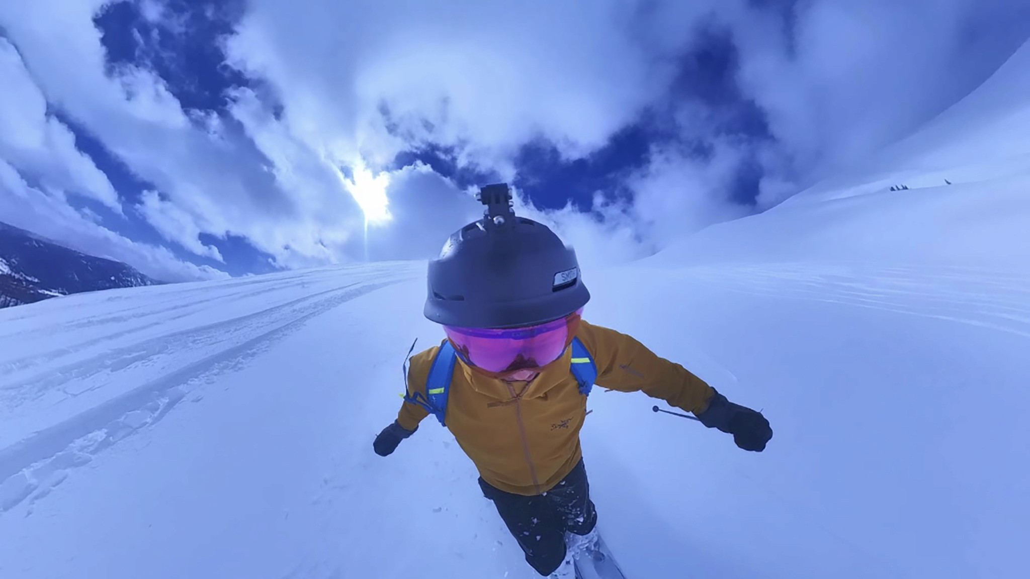 A person skiing downhill on a snowy mountain, wearing a yellow jacket, black pants, and a helmet with a camera, under a bright blue sky with scattered clouds.