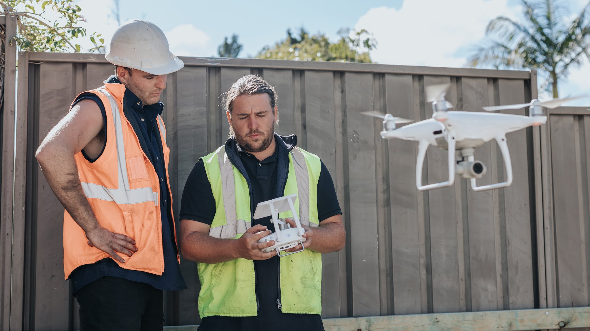 Two construction workers in safety vests and helmets operate a white drone in front of a wooden fence on a sunny day, focusing on the drone's remote controller.
