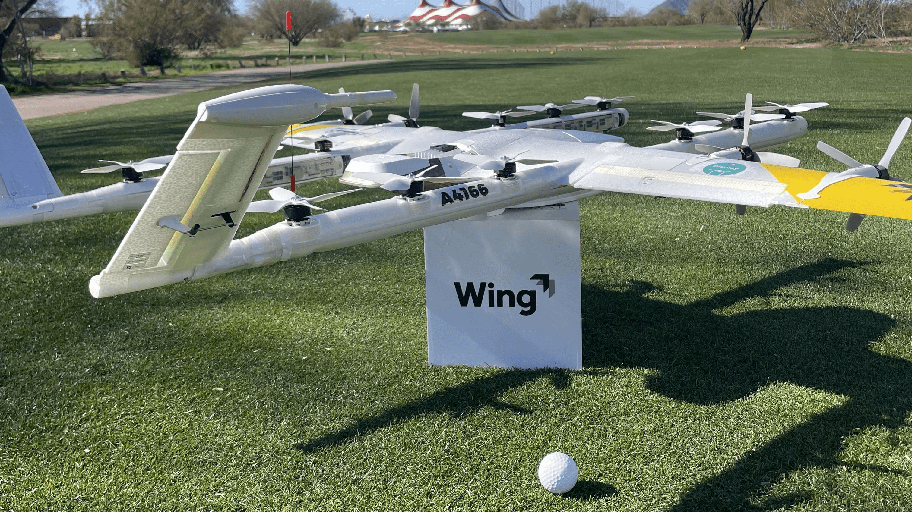 A delivery drone with multiple rotors is showcased on a grassy field near a golf ball, featuring the brand "Wing" prominently displayed in front of it.