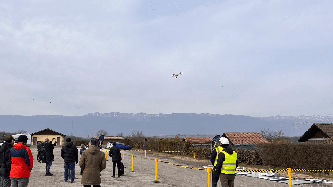 A group of people stand on an open airfield watching a small aircraft take flight against a backdrop of overcast skies and distant mountains, with some wearing winter clothing and one person in a high-visibility vest near a caution barrier.