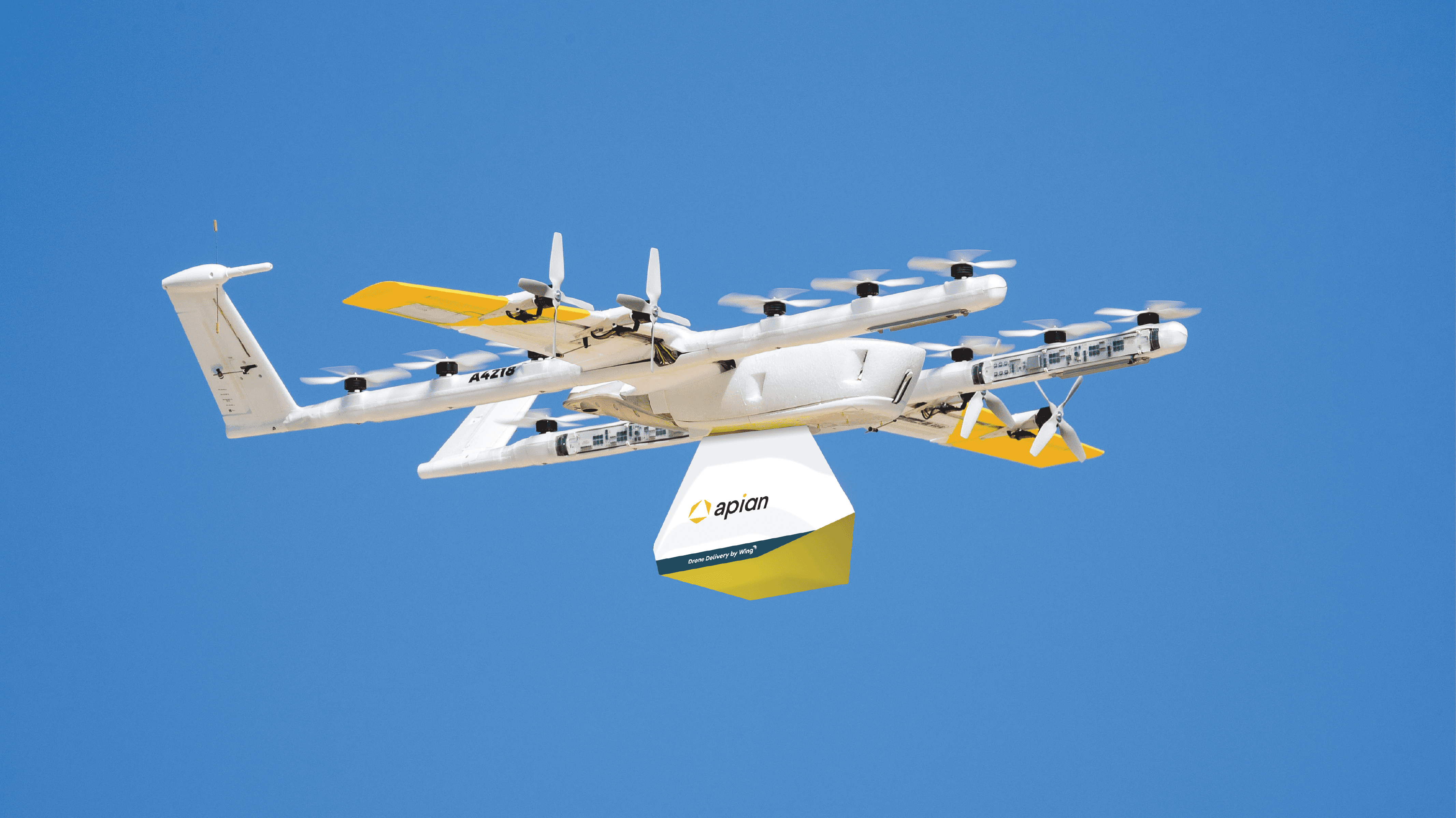 A white and yellow drone in flight against a clear blue sky; it is carrying a branded Apion package, demonstrating modern delivery technology.