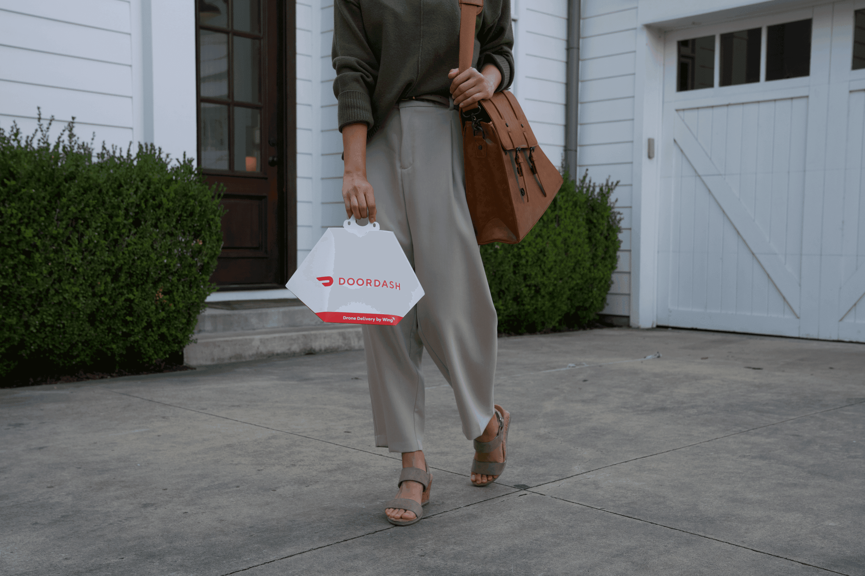 A person carrying a DoorDash takeout bag walks on a concrete driveway, wearing beige pants, brown sandals, and carrying a brown handbag, with a white house and garage in the background.
