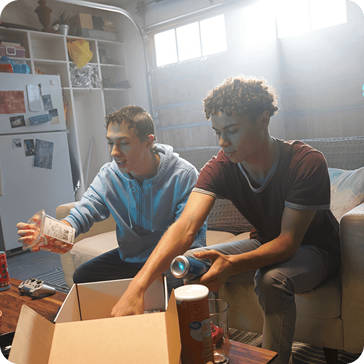 Two young people in a well-lit room, characterized by cozy decor and filled bookshelves, sort through a cardboard box containing various items, including snacks, creating a relaxed and casual atmosphere.