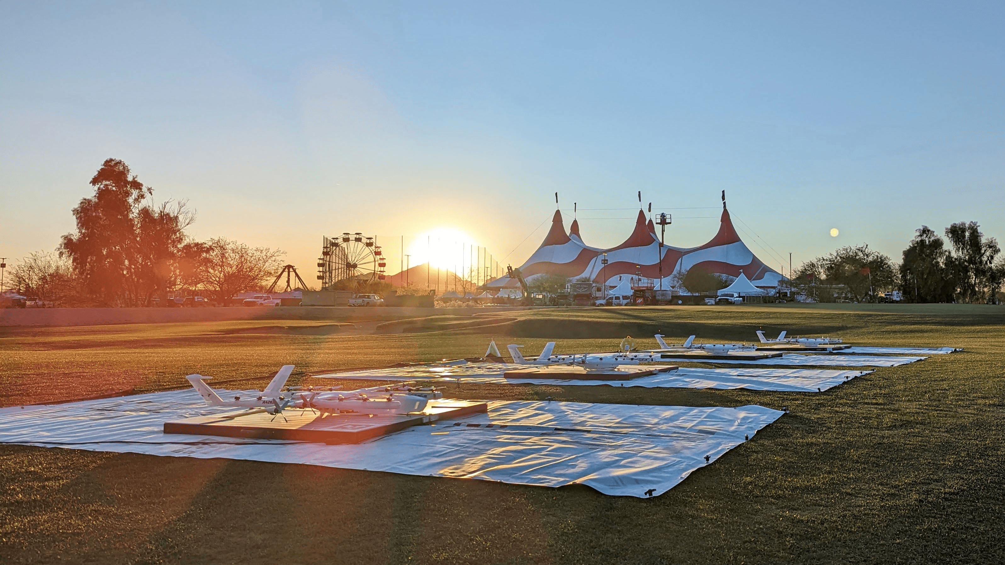 Sun setting behind a field with small planes on tarps, adjacent to a circus tent, with a clear sky and trees in the background.